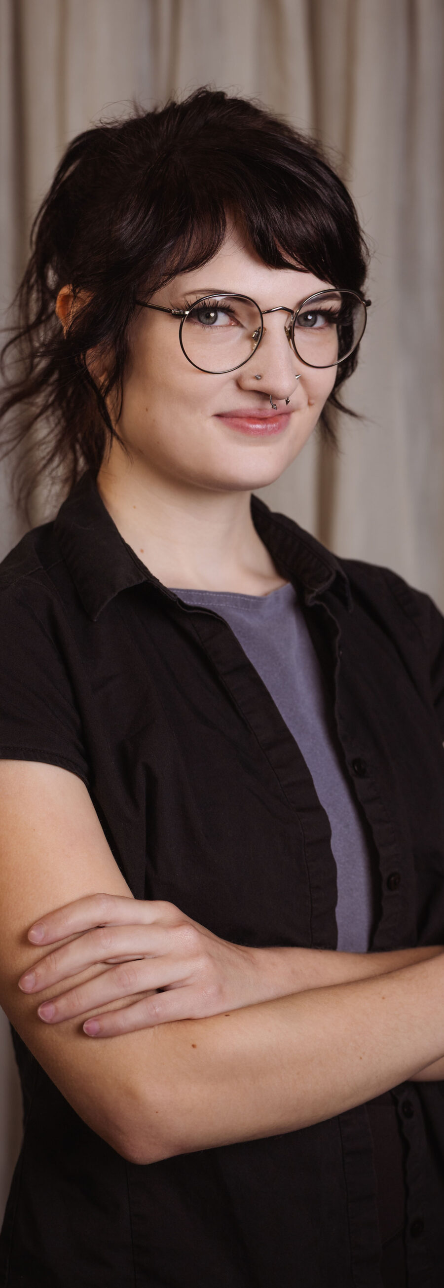 Grace, smiling with arms crossed, wearing glasses and a black shirt in front of a studio curtain backdrop