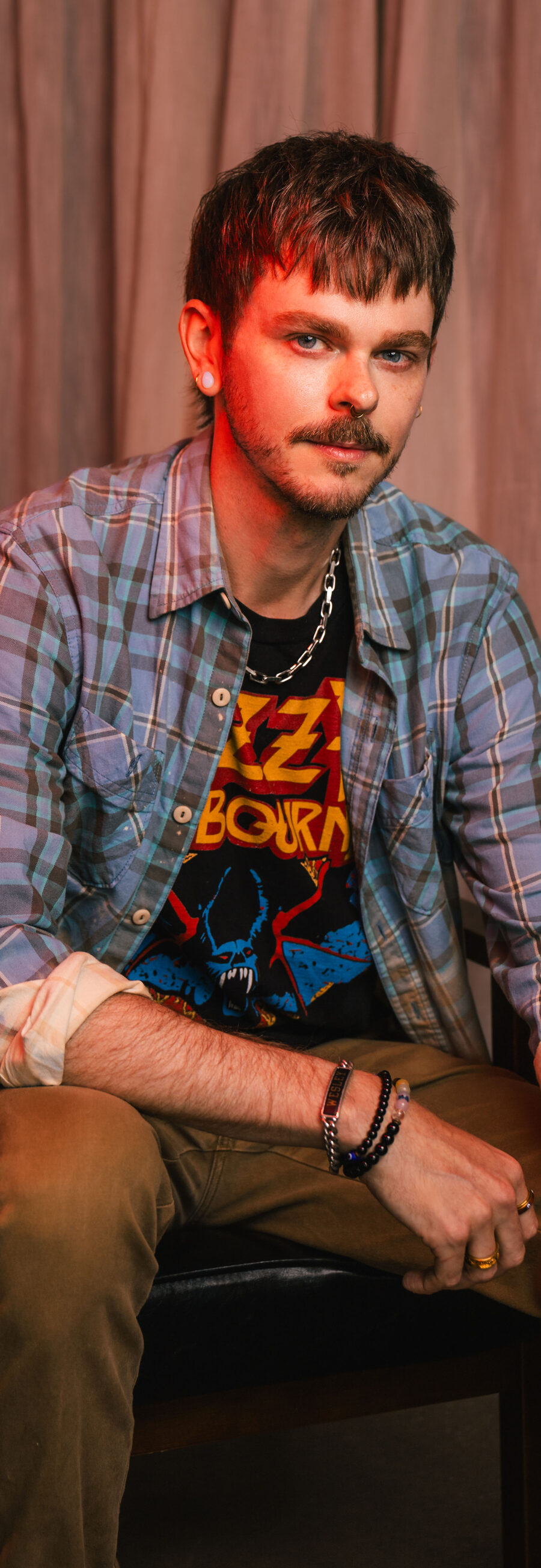 Wes, seated in a leather chair, wearing a plaid shirt over a band tee, in front of a studio curtain backdrop